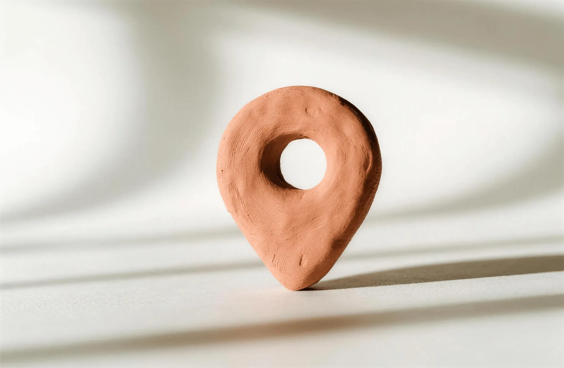 Een met de hand gekneed locatiespeldje van terracottakleurige klei, staand op een lichte ondergrond in zacht daglicht.