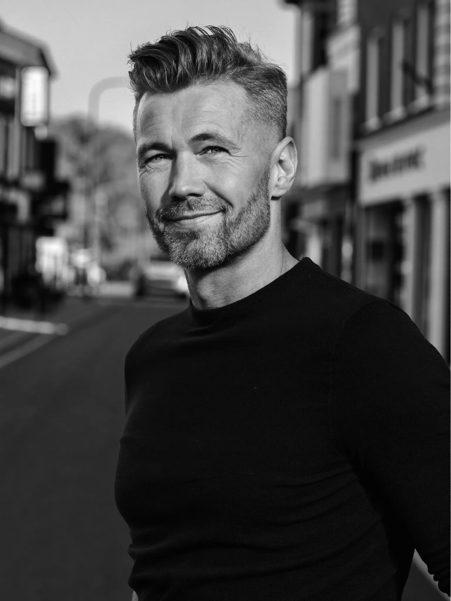 Klassiek heren — Zwart-witportret van een man van rond de vijftig met een klassiek kort kapsel en nette scheiding. Onscherpe achtergrond van een Nederlandse straat.