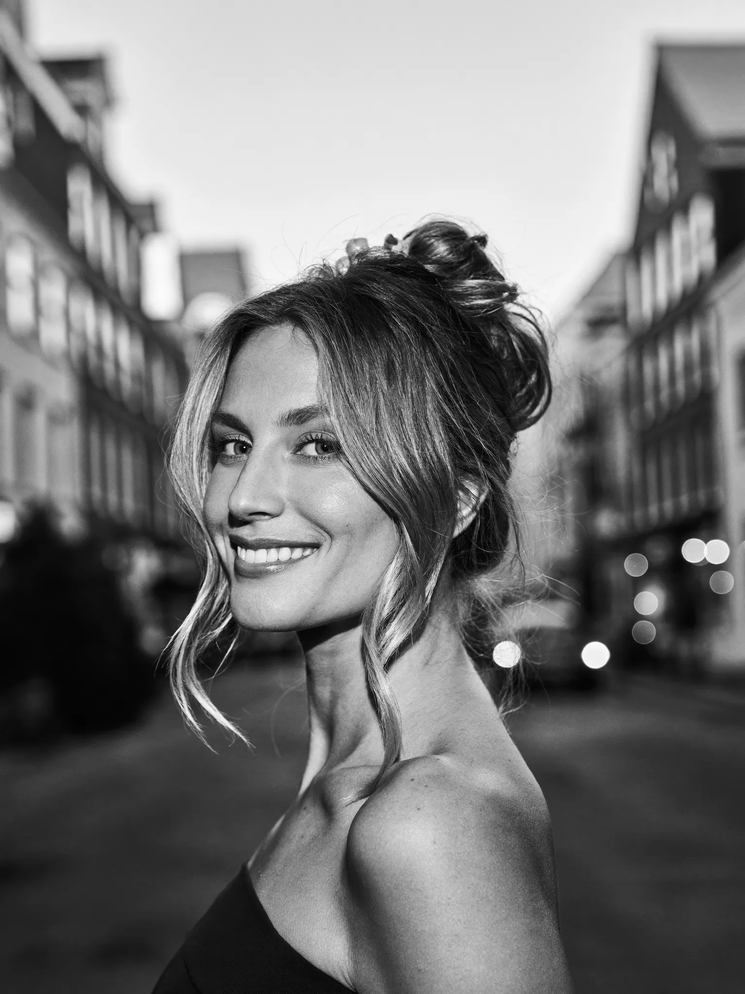 Opsteken — Zwart-witportret van een vrouw met feestelijk opgestoken haar, half van achteren zodat de vorm van het kapsel zichtbaar is. Onscherpe avondstraat op de achtergrond.